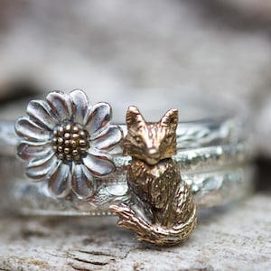Puede incluir: Un anillo de plata con una margarita y un dije de zorro dorado. La margarita tiene pétalos plateados y un centro marrón. El zorro está detallado y sentado. La banda del anillo tiene un diseño texturizado.