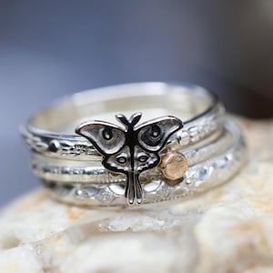 May include: A stack of silver rings with intricate designs. The top ring features a detailed silver moth charm. A small gold accent is visible on one of the rings. The rings are displayed on a textured surface.