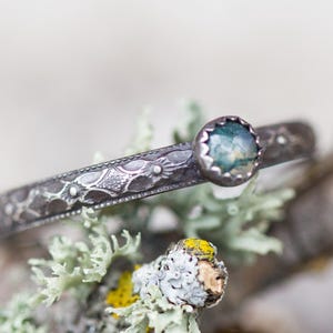 Könnte beinhalten: Ein silbernes Armband mit einem eingravierten Blumenmuster und einem runden, eingefassten Edelstein. Der Edelstein hat einen blau-grünen Farbton. Das Armband wird auf einem Ast mit Flechten präsentiert.