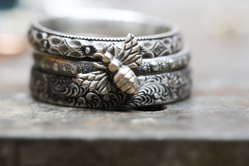 May include: A stack of three silver rings with intricate floral and geometric patterns. A detailed bee charm is attached to the top ring. The rings are displayed on a textured surface, showcasing the craftsmanship of the jewelry.
