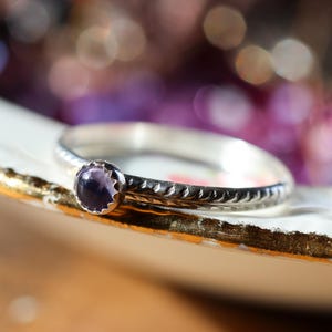 May include: A silver ring with a small, round, purple gemstone set in a decorative bezel. The band features an etched design. The ring is displayed on a white and gold-trimmed surface, with a blurred background of purple and gold.
