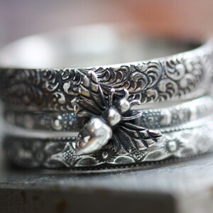 May include: A stack of three silver rings with intricate floral and dotted patterns. A detailed bee charm is centered on the top ring. The rings have a vintage, handcrafted appearance.