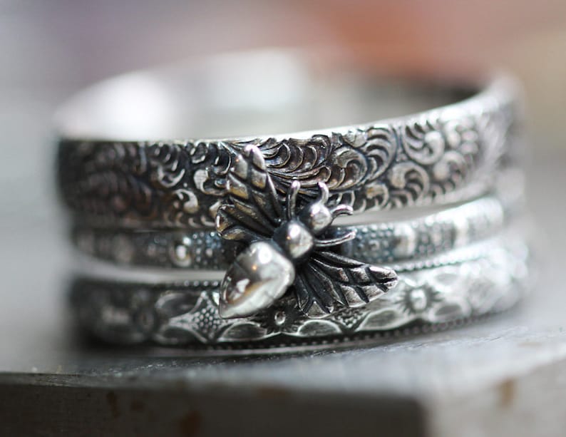 May include: A stack of three silver rings with intricate floral and dotted patterns. A detailed bee charm is centered on the top ring. The rings have a vintage, handcrafted appearance.
