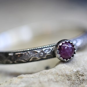 Pu&ograve; includere: Un anello d'argento con un designo ornato e una piccola pietra preziosa rotonda di colore rosa.