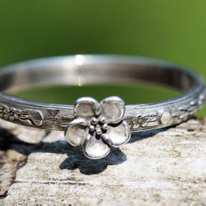 May include: A silver-toned metal ring featuring a band with an embossed floral pattern and a prominent five-petaled flower ornament centered on the front. The metal surface has a slightly oxidized finish that highlights the intricate details.