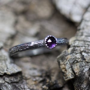 May include: A silver ring with a purple gemstone set in a bezel setting. The ring band is decorated with a floral design.