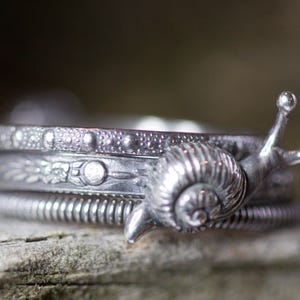 Può includere: Un braccialetto d'argento con un ciondolo a forma di lumaca, composto da tre bande, ciascuna con un design diverso. La banda superiore ha una superficie testurizzata, la banda centrale ha un motivo floreale e la banda inferiore è un semplice filo ritorto.