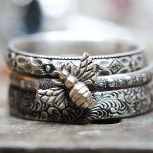 May include: A stack of three silver rings with intricate floral and geometric patterns. A detailed silver bee charm is attached to the rings. The rings are stacked on top of each other.