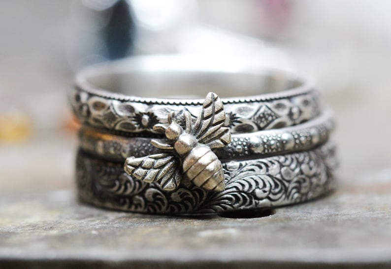 May include: A stack of three silver rings with intricate floral and geometric patterns. A detailed silver bee charm is attached to the rings. The rings are stacked on top of each other.