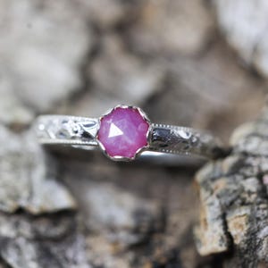 May include: A silver ring with a pink gemstone set in a faceted bezel. The ring band has an intricate engraved design.