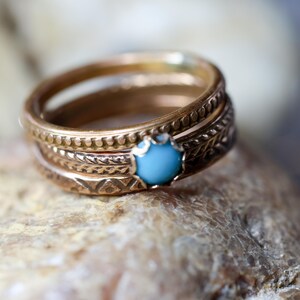 May include: A stack of gold-colored rings with intricate embossed patterns. One ring features a round, light blue gemstone. The rings are stacked on a textured, light brown surface.