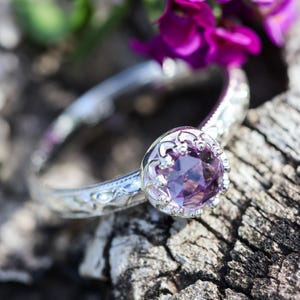 May include: A silver ring featuring a round, faceted purple gemstone set in a crown-like prong setting. The band of the ring displays an intricate, embossed floral pattern along its surface, providing a detailed texture to the metallic band.