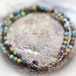 May include: A handcrafted bracelet featuring two strands of colorful beads in shades of blue, green, brown, and yellow. The bracelet is secured with a gold-colored clasp and is resting on a textured stone surface. The beads appear to be approximately 0.25 inches in diameter.