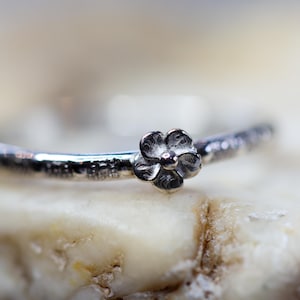 Puede incluir: Anillo de plata con un pequeño diseño floral detallado. La banda del anillo tiene una apariencia texturizada. La flor, centrada, tiene un acabado oscuro y oxidado, creando contraste con la banda pulida.