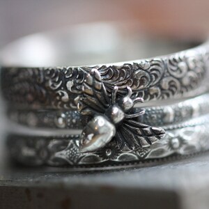 May include: A stack of three silver rings with intricate floral and scrollwork patterns. The top ring features a detailed bee charm with a pear-shaped body and delicate wings. The rings are stacked, creating a layered effect.