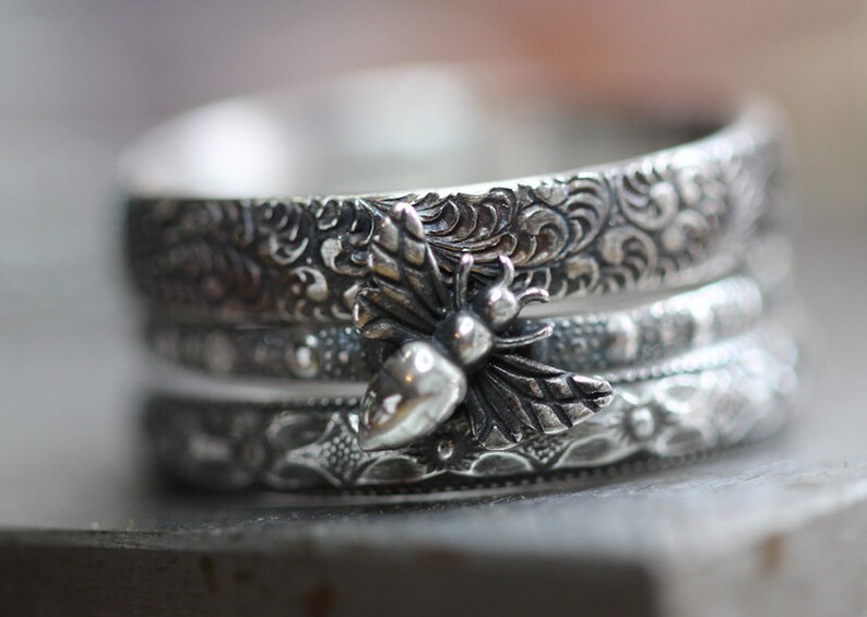 May include: A stack of three silver rings with intricate floral and scrollwork patterns. The top ring features a detailed bee charm with a pear-shaped body and delicate wings. The rings are stacked, creating a layered effect.