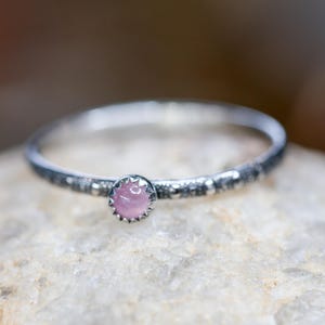 May include: A silver ring with a small, round, pink gemstone. The ring band has a textured design. The gemstone is set in a decorative bezel. The ring is displayed on a light-colored, textured surface.