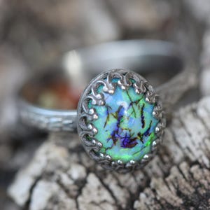 May include: A silver ring featuring an oval-shaped stone with a light green and blue iridescent surface, accented by dark purple and black inclusions. The stone is set in a decorative, scalloped metal bezel with a textured, patterned band.