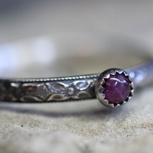 Pu&ograve; includere: Un anello d'argento con un designo ornato e una singola pietra preziosa rotonda di colore viola.