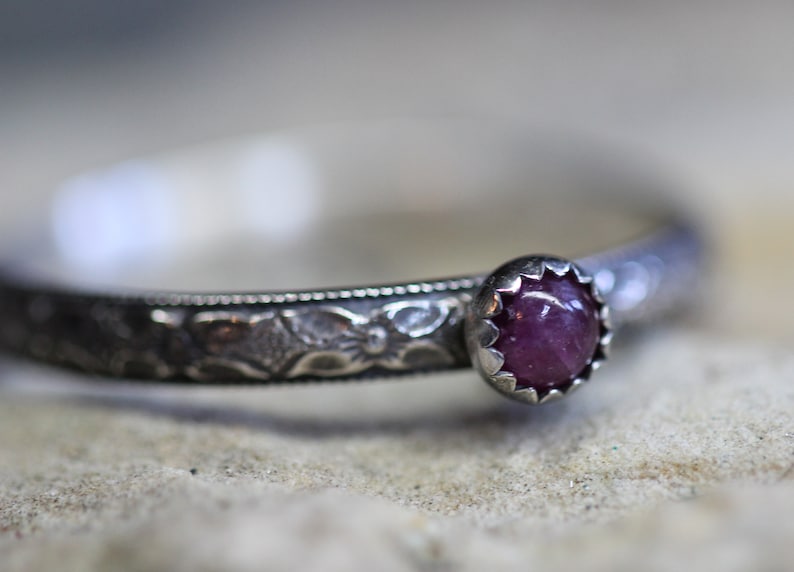 Pu&ograve; includere: Un anello d'argento con un designo ornato e una singola pietra preziosa rotonda di colore viola.