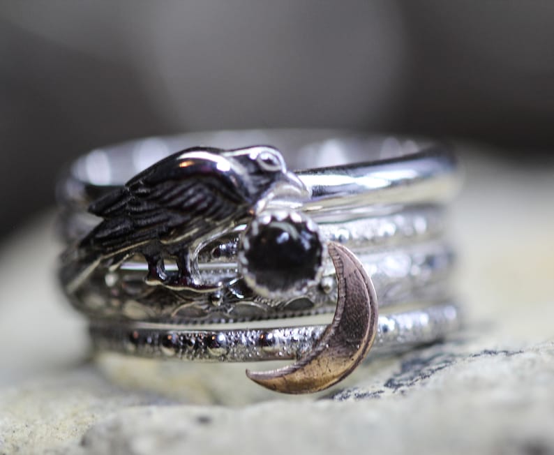 May include: A set of five silver rings with a detailed design. The top ring features a black raven with a black gemstone. The second ring has a crescent moon design. The remaining rings have a variety of textured patterns.