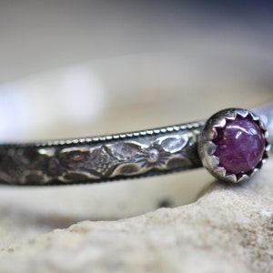 May include: A silver ring with an ornate band and a single pink gemstone set in a silver bezel.
