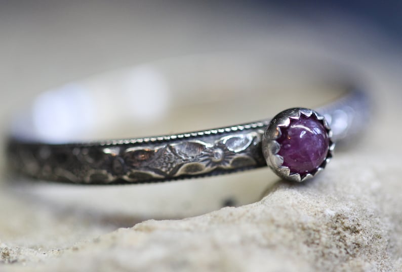 Pu&ograve; includere: Un anello d'argento con una fascia decorata e una singola pietra preziosa rosa incastonata in una montatura d'argento.