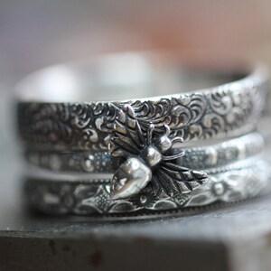 May include: A stack of three silver rings with intricate floral and scrollwork patterns. The top ring features a detailed bee charm with wings and a rounded body, adding a nature-inspired touch to the jewelry.