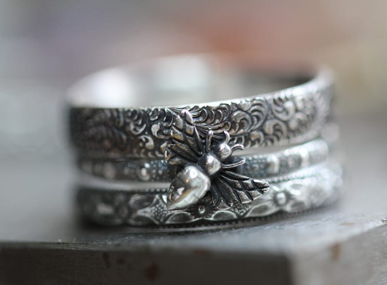 May include: A stack of three silver rings with intricate floral and scrollwork patterns. The top ring features a detailed bee charm with wings and a rounded body, adding a nature-inspired touch to the jewelry.