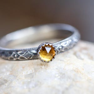 May include: A silver ring with an engraved band and a round, amber-coloured gemstone. The ring is set on a white stone surface, with a blurred background. The gemstone is the focal point of the ring.