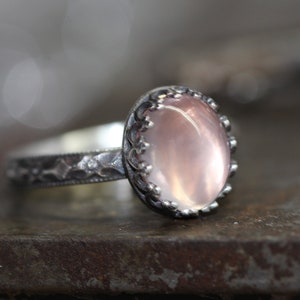 May include: A silver ring featuring a delicate floral design and a pale pink oval gemstone.