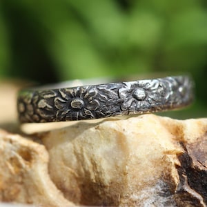 Può includere: Un anello a fascia d'argento con un designo floreale. L'anello è in argento sterling e presenta un delicato motivo floreale.