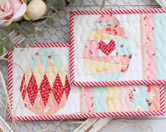 Könnte beinhalten: Zwei gequiltete Topflappen in Form von Ostereiern mit roten und weißen Streifen an den Rändern. Die Topflappen sind aus Patchwork-Stoff in den Farben Rosa, Gelb und Blau gefertigt.