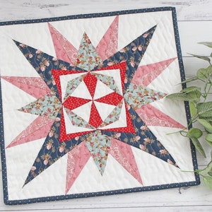May include: A small, quilted square with a starburst design in shades of pink, red, blue, and white. The starburst is surrounded by a navy blue border with white polka dots.