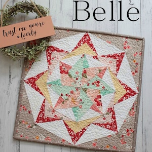 May include: A beige and brown quilted square with a multi-coloured star design in the centre. The star is made of red, yellow, pink, and green fabric. The text "Belle" is written in black above the square.