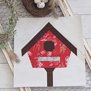 Könnte beinhalten: Ein roter und brauner Vogelhaus-Patchworkblock mit einem Blumenmuster. Das Vogelhaus hat ein weißes Dach und eine braune Tür.