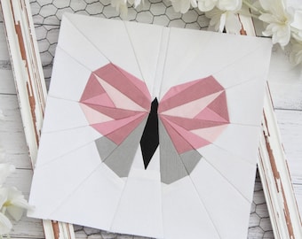 May include: A white fabric square with a pink and gray butterfly design. The butterfly is made of fabric pieces sewn together to create a geometric pattern.