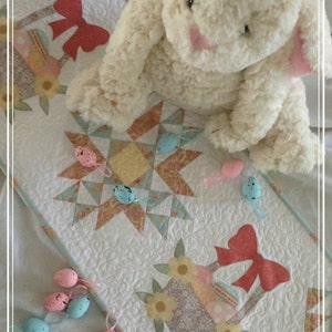 May include: A white quilted table runner with a pastel blue border features a patchwork design with a star in the centre. The runner has two appliqued baskets with bows and flowers. There are several small pastel blue and pink eggs scattered around the runner.
