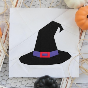 May include: A white fabric square with a black witch's hat featuring a purple and red band. The hat is set against a white background with a white frame surrounding it. There are pumpkins and other Halloween decorations around the frame.