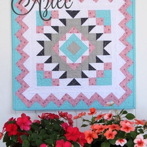 May include: A colorful quilt with a geometric pattern in shades of pink, turquoise, gray, and black. The quilt is titled "Aztec" and is hanging on a white wall. Two potted plants with pink and orange flowers are in the foreground.
