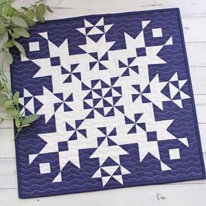 Puede incluir: Textil acolchado cuadrado con un patrón geométrico de formas blancas sobre un fondo azul marino. El diseño incluye motivos de estrellas y molinetes. Los bordes están acabados con un borde azul marino y una puntada ondulada.