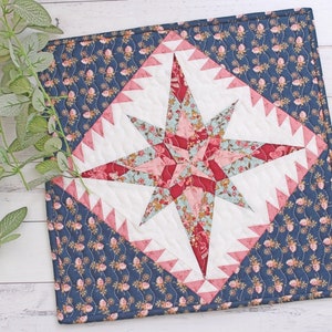 May include: A quilted patchwork star design with a white background and pink, red, and blue fabric. The star is surrounded by a navy blue fabric with a floral pattern.