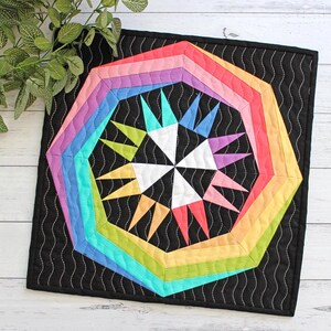 May include: A colorful, geometric quilt with a rainbow design. The quilt features a central starburst pattern in white, surrounded by a series of rainbow-colored octagons. The background is black with a wavy white stitch pattern. The quilt is square.