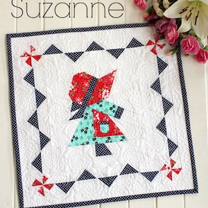 May include: A white quilt block with a red, blue, and green patchwork design of a girl in a dress. The quilt block is surrounded by a navy blue and white polka dot border. The text "Suzanne" is at the top of the image and "includes bonus mirror image FPP pattern" is at the bottom.