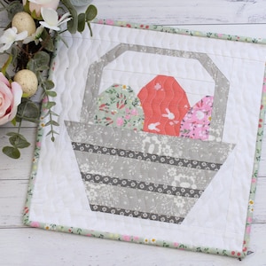 May include: A white quilted square with a green border features a gray and white basket filled with three Easter eggs. The eggs are decorated with floral patterns and bunny silhouettes. One egg is pink, one is green, and one is red.