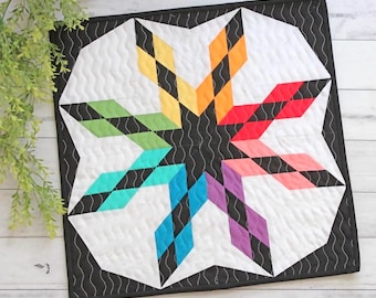May include: A vibrant, geometric quilt with a starburst pattern. The quilt has a white backdrop, a black border, and a spectrum of colours: yellow, orange, red, pink, purple, blue, and green. The quilt measures approximately 30 cm square.