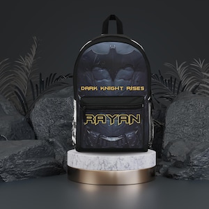 May include: Black backpack with a Batman graphic and the text "DARK KNIGHT RISES". The backpack has a yellow zipper and the name "RAYAN" is written in yellow letters on the front.