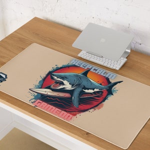 May include: A beige desk mat with a graphic of a shark riding a surfboard in front of a red and orange sun. The text "Keep Calm Game On" is printed on the mat.