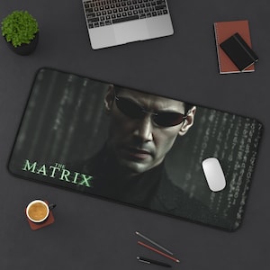 May include: A black rectangular mousepad featuring an image of a man wearing sunglasses, with the text "THE MATRIX" in green. The background has a digital rain effect. A white mouse is on the pad.