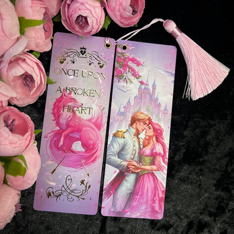 Peut inclure: Deux marque-pages d&eacute;coratifs aux tons roses et violets. L'un pr&eacute;sente un renard rose et le texte "Once Upon a Broken Heart". L'autre repr&eacute;sente un couple devant un ch&acirc;teau. Une pompon rose est attach&eacute;e en haut.